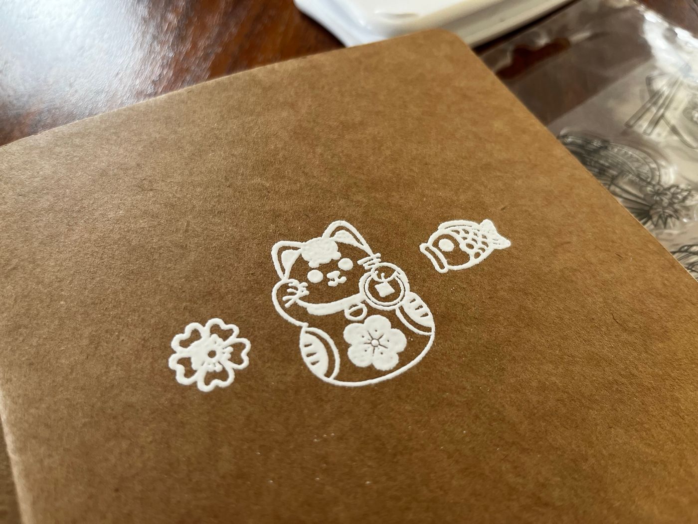 Eine weiße Relief-Prägung japanischer Motive wie Koi und Kirschblüte auf dem braunen Cover eines Kraftpapier-Notizbuchs.
