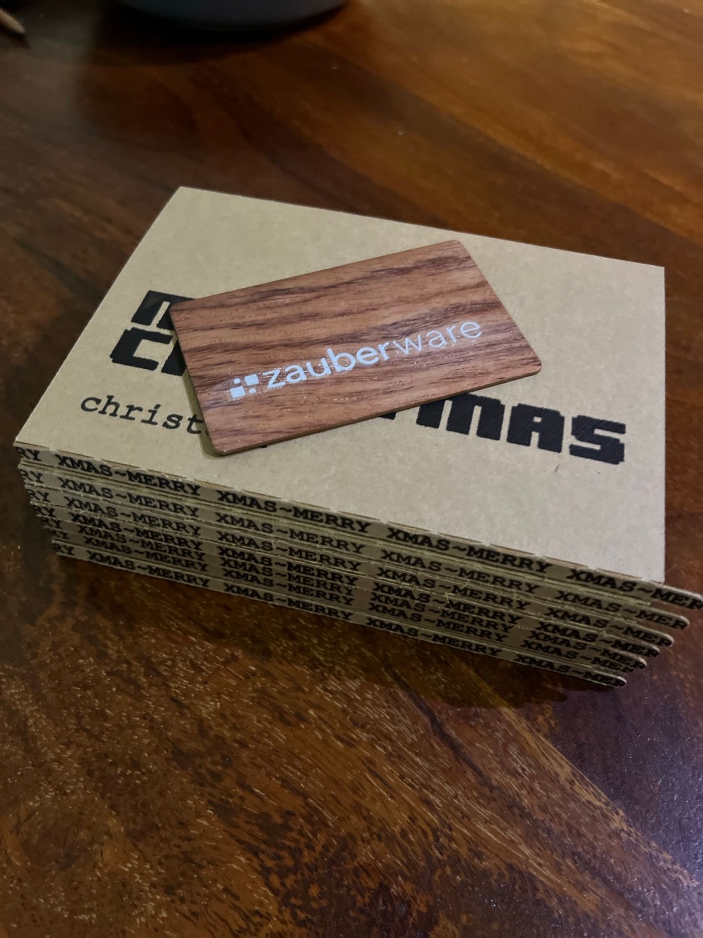 Eine NFC-Visitenkarte aus dunklem Nussbaumholz mit weißem Logo liegt oben auf einem Stapel handgefertigter Geschenkverpackungen.