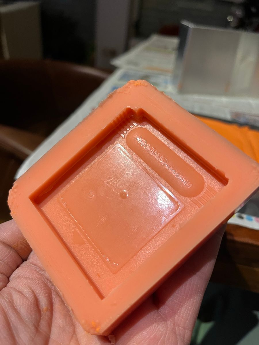 Eine Hand hält eine fertig gegossene orangefarbene Silikonform mit dem detailreichen Negativ-Abdruck einer Game-Boy-Spielekassette.